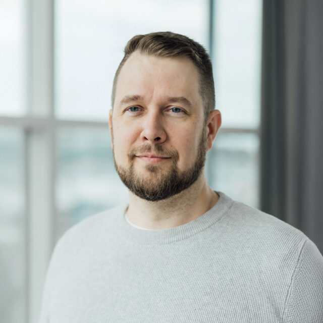 Mies seisoo sisätilassa ikkunan edessä, yllään vaalea neulepaita. Hänellä on parta ja lyhyet hiukset. Taustalla näkyy sumeaa maisemaa ikkunan läpi.